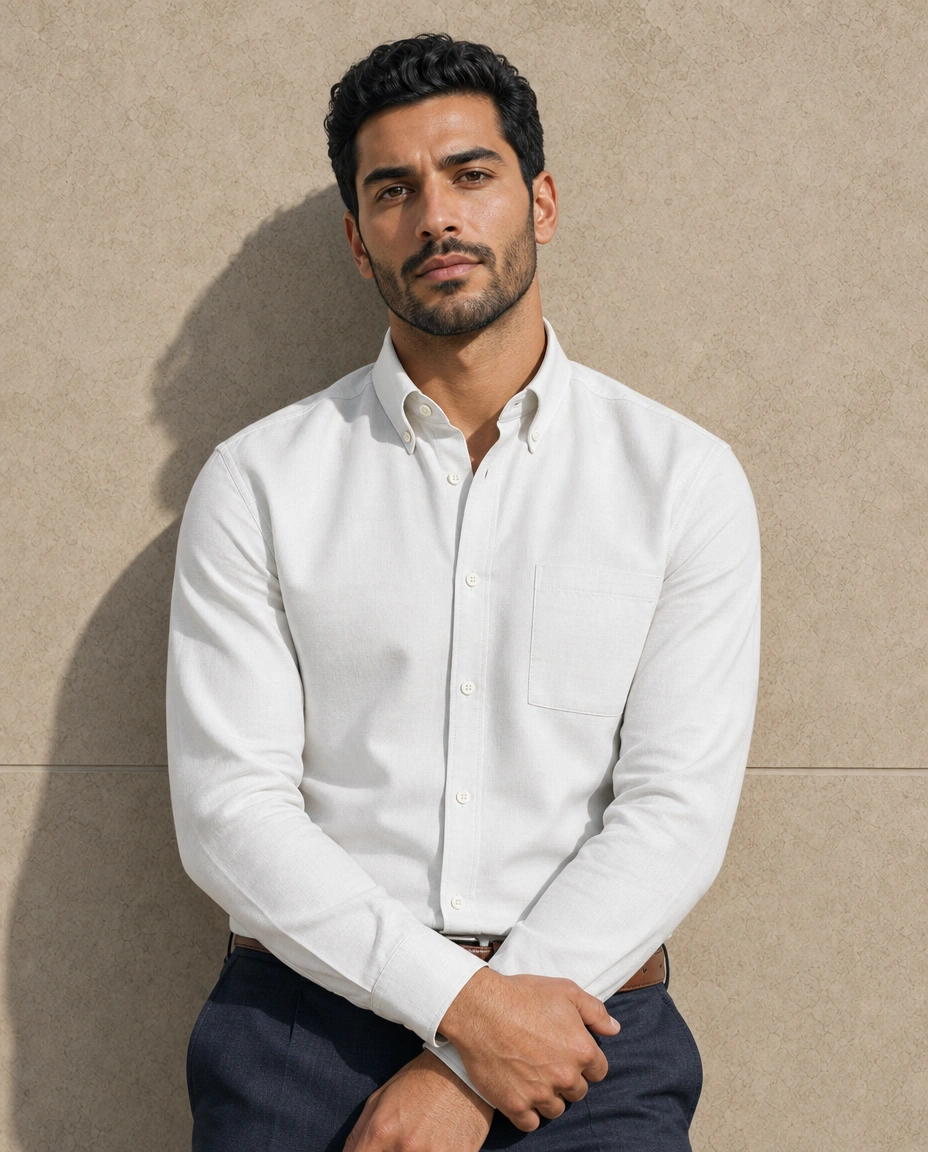 A model against a sunlit stone wall in a cream long-sleeved button-down shirt with a chest pocket and deep blue trousers
