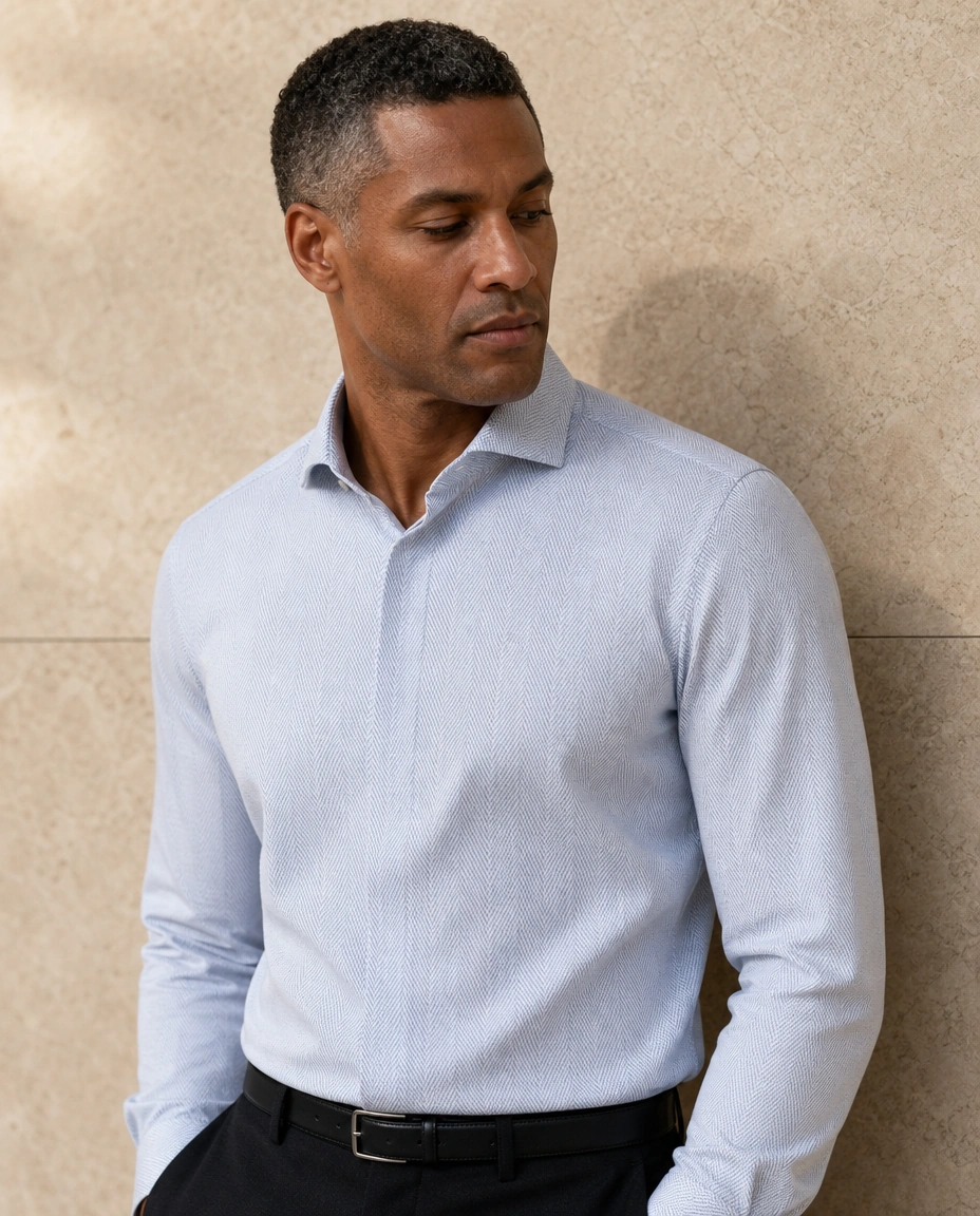 A model against a sunlit stone wall in a pale blue long-sleeved shirt with a cutaway collar and soft black trousers
