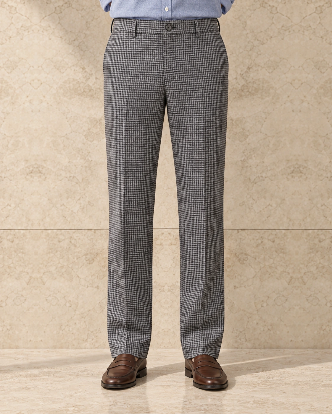 A model from the waist down against a sunlit stone wall, in light grey houndstooth trousers on a single-button waistband, flat-fronted, high rise and straight-legged over brown loafers