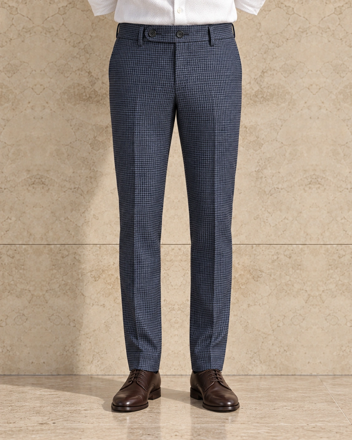 A model from the waist down against a sunlit stone wall, in airforce blue houndstooth trousers on an extended tab with two buttons, flat-fronted, mid rise and slim tapered over brown lace-ups