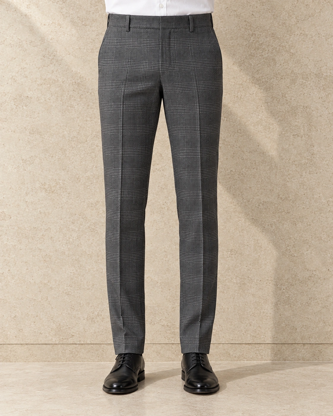 A model from the waist down against a sunlit stone wall, in mid grey glen check trousers on a plain waistband closed with a hook and bar, flat-fronted and hemmed plain
