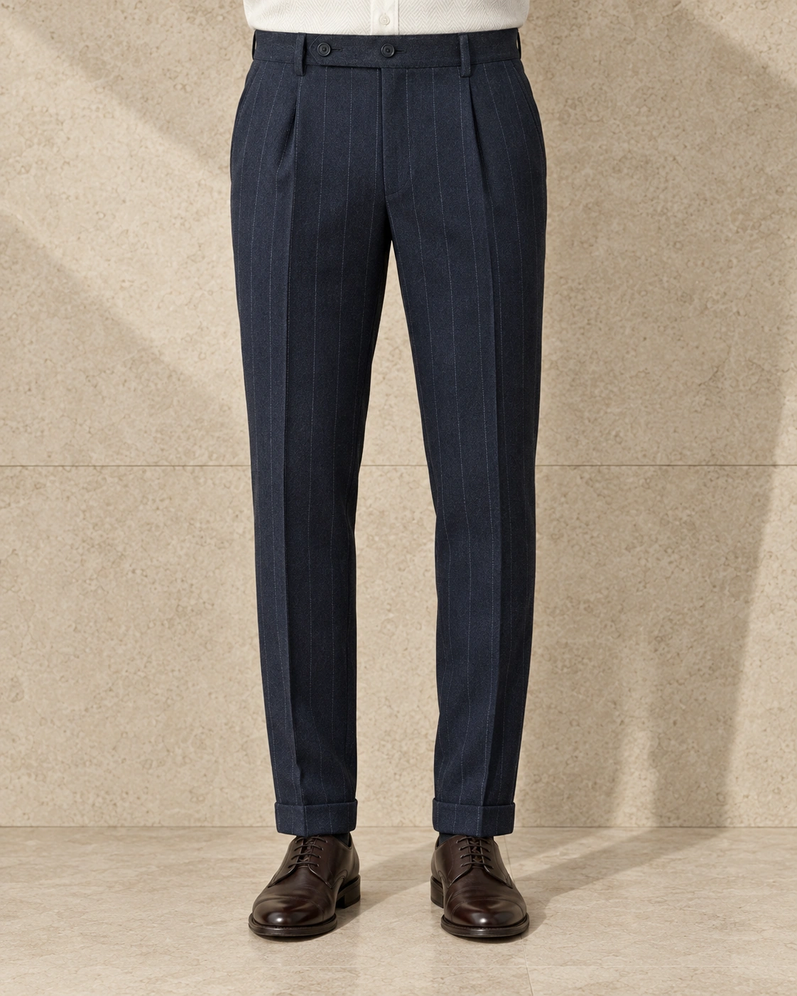 A model from the waist down against a sunlit stone wall, in navy chalk stripe trousers on an extended tab with two buttons, single-pleated and finished with turn-ups over brown lace-ups