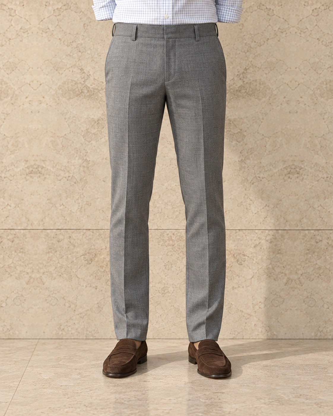 A model from the waist down against a sunlit stone wall, in light grey trousers on a plain waistband closed with a hook and bar, flat-fronted, low rise and slim tapered over brown suede loafers