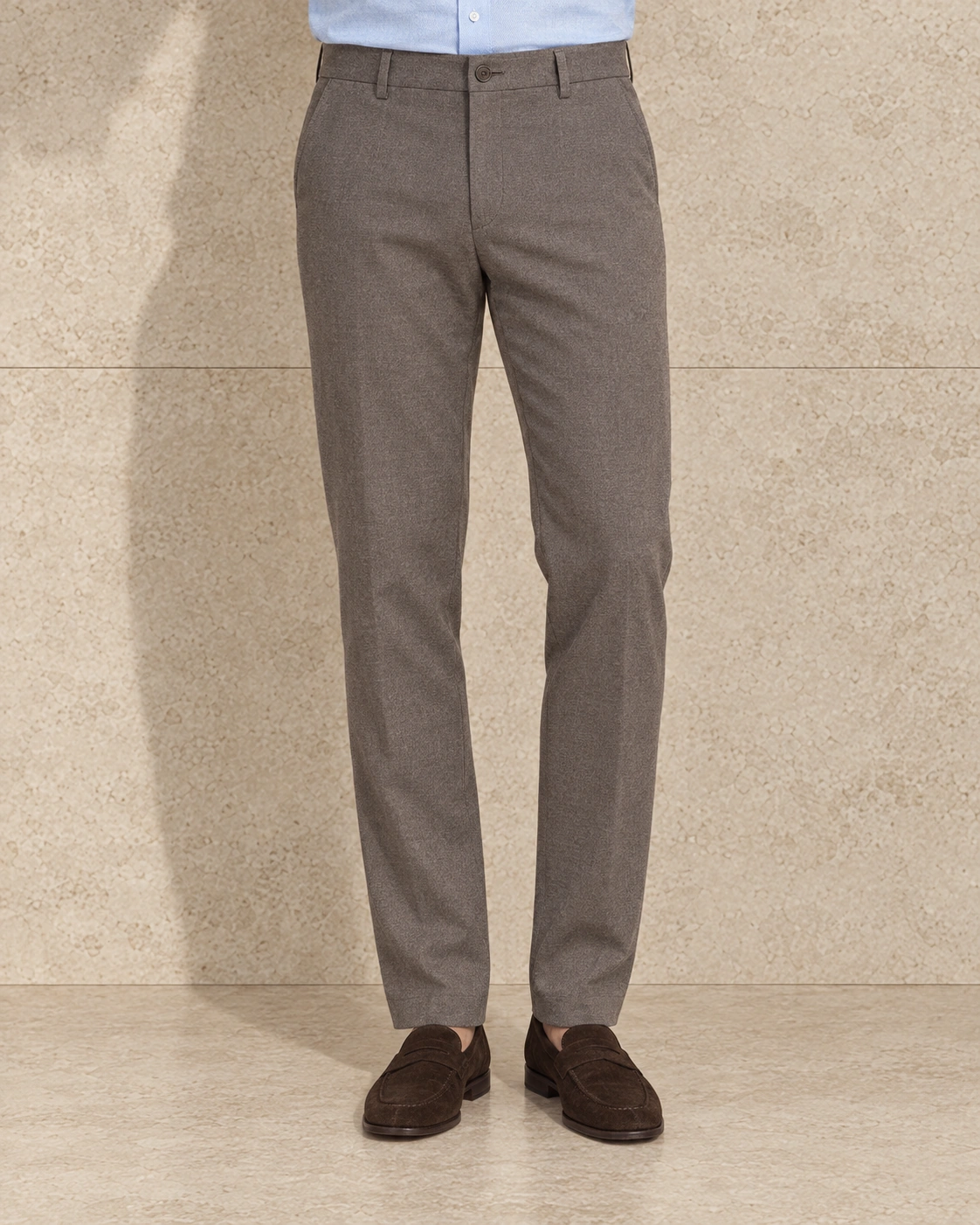 A model from the waist down against a sunlit stone wall, in light taupe trousers on a single-button waistband, flat-fronted and hemmed plain over brown suede loafers