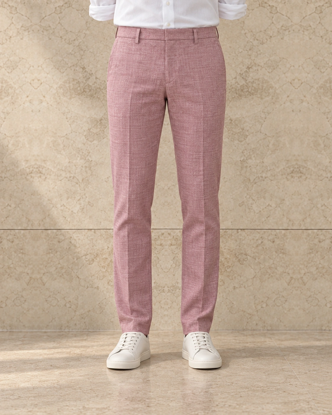 A model from the waist down against a sunlit stone wall, in rose trousers on a plain waistband closed with a hook and bar, flat-fronted, mid rise and slim tapered over white sneakers