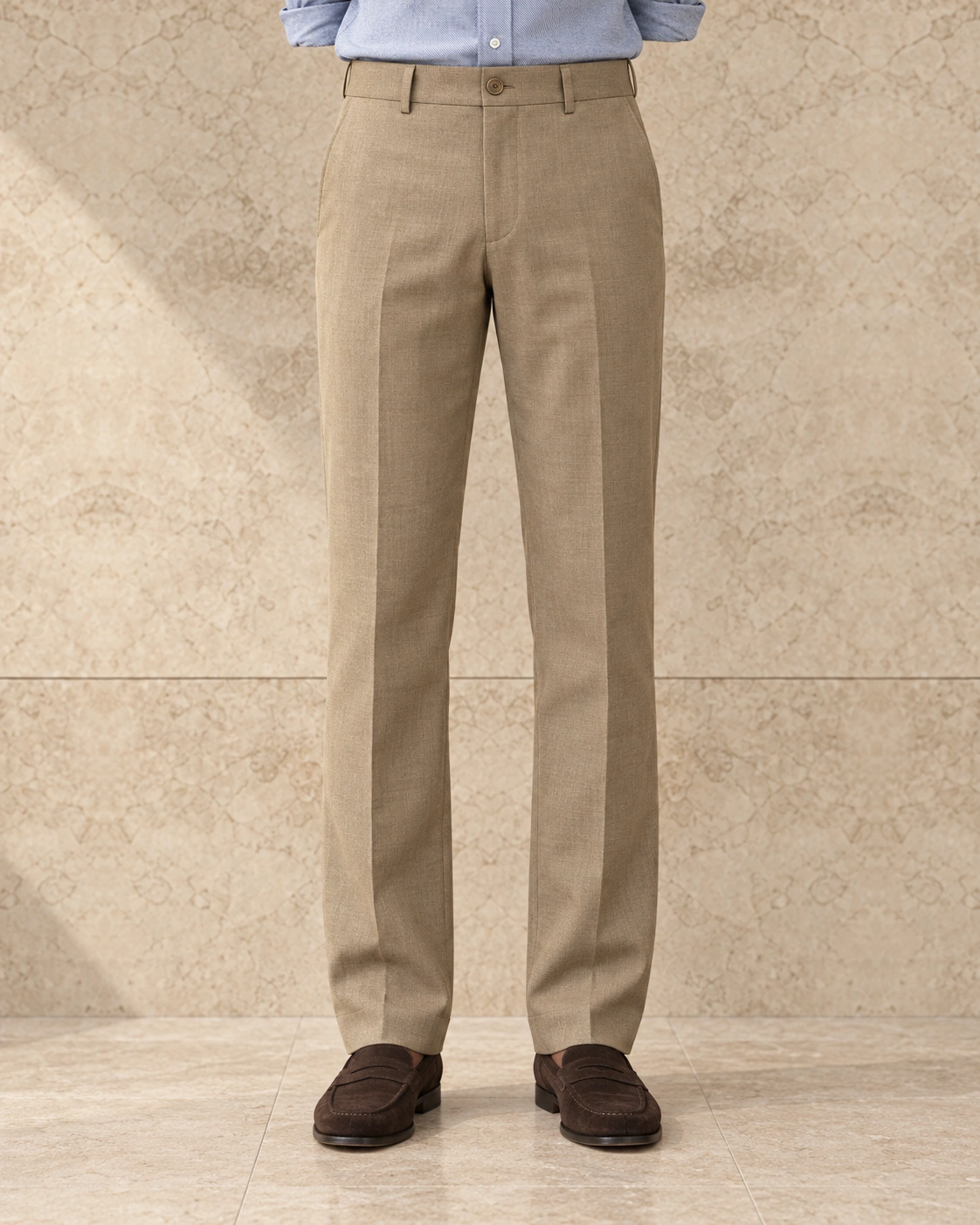 A model from the waist down against a sunlit stone wall, in camel trousers on a single-button waistband, flat-fronted, high rise and straight-legged over brown suede loafers