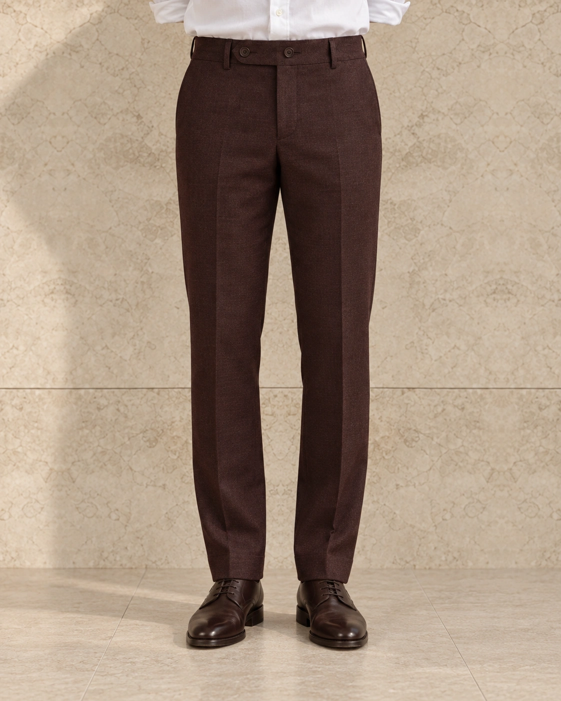 A model from the waist down against a sunlit stone wall, in chestnut trousers on an extended tab with two buttons, flat-fronted, mid rise and tapered over dark brown lace-ups