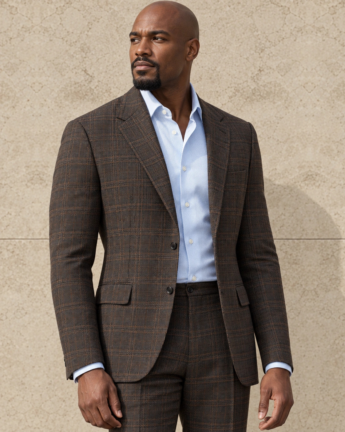 A model against a sunlit stone wall in a soft-shouldered brown check two-piece suit, worn open-necked over a pale blue shirt